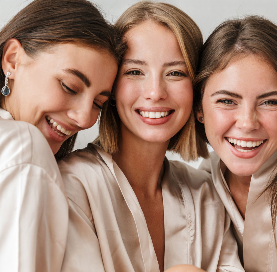 Three women smiling in satin robes, models with smooth glowing skin